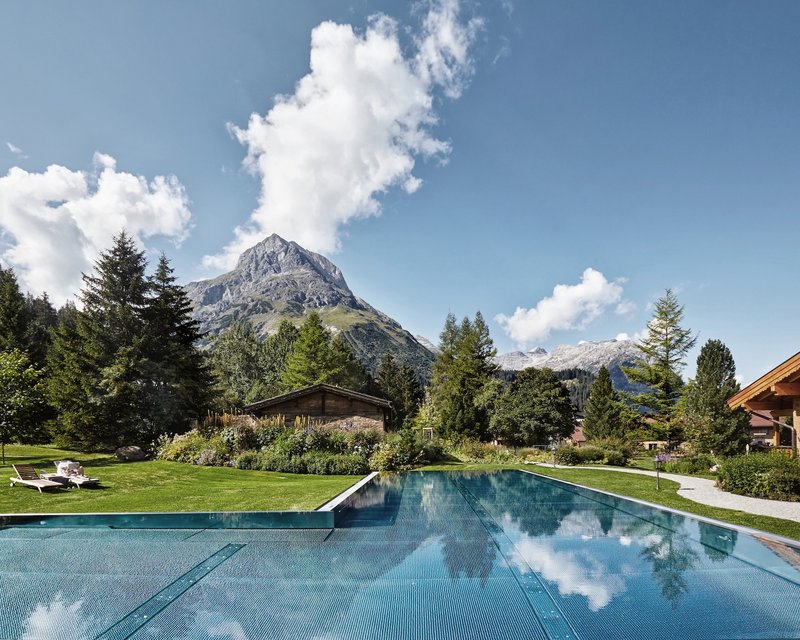 Neues aus dem Hotel Post Lech Schwimmbad vor Bergen und Bäumen unter blauem Himmel mit Wolken