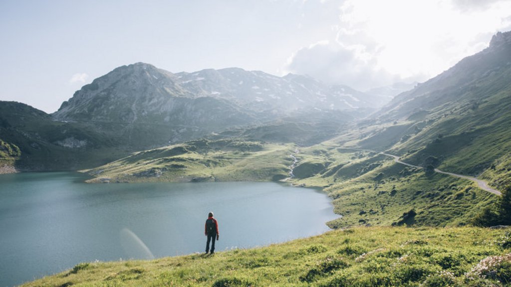 Sommer am Arlberg | Alpine Natur, Bewegung & Erholung im Hotel Post Lech Person in roter Jacke blickt auf Bergsee und Berge bei bewölktem Himmel